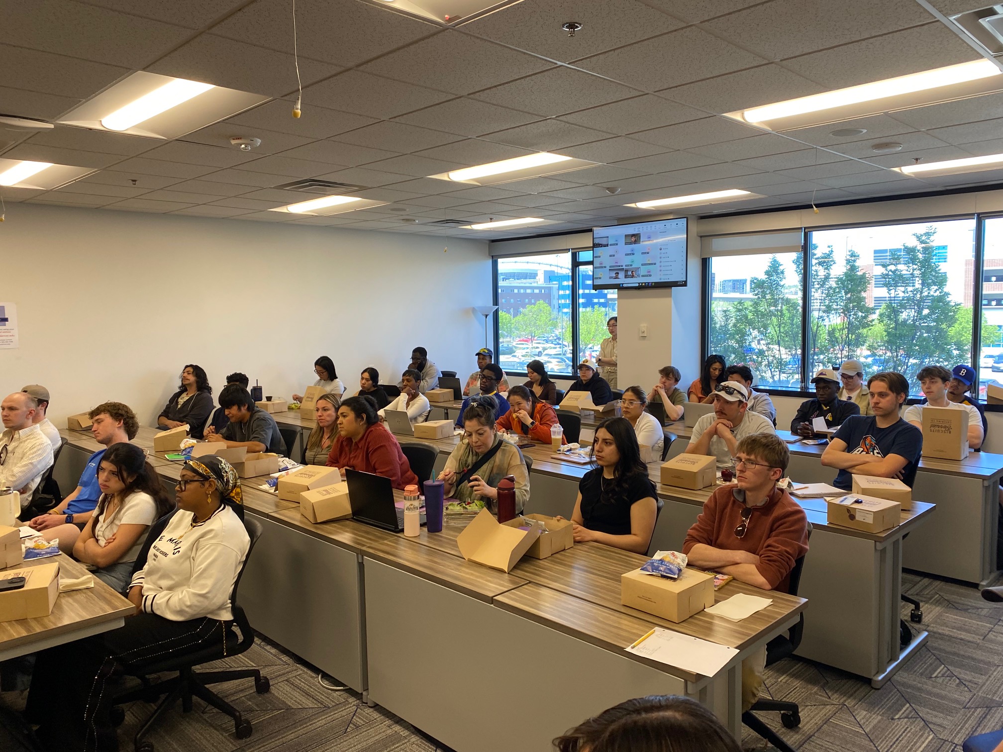 A wide-angle shot of a crowded classroom or training room filled with young adults seated at long desks. Most students are looking toward the front of the room, and many have cardboard lunch boxes and water bottles in front of them. In the background, large windows show a bright outdoor view of buildings and trees, and a flat-screen monitor is mounted on the wall displaying a video conference interface.