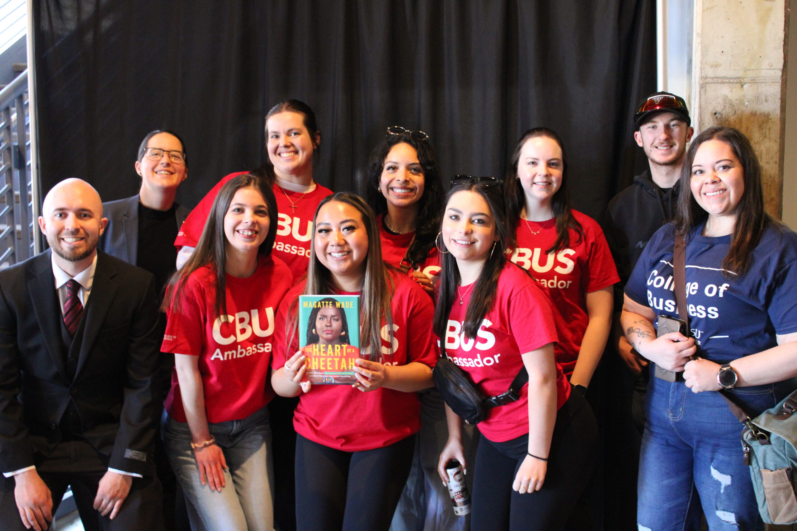 A group of students and a professional-looking man in a suit pose together in front of a black curtain. Most of the students are wearing red t-shirts that say 