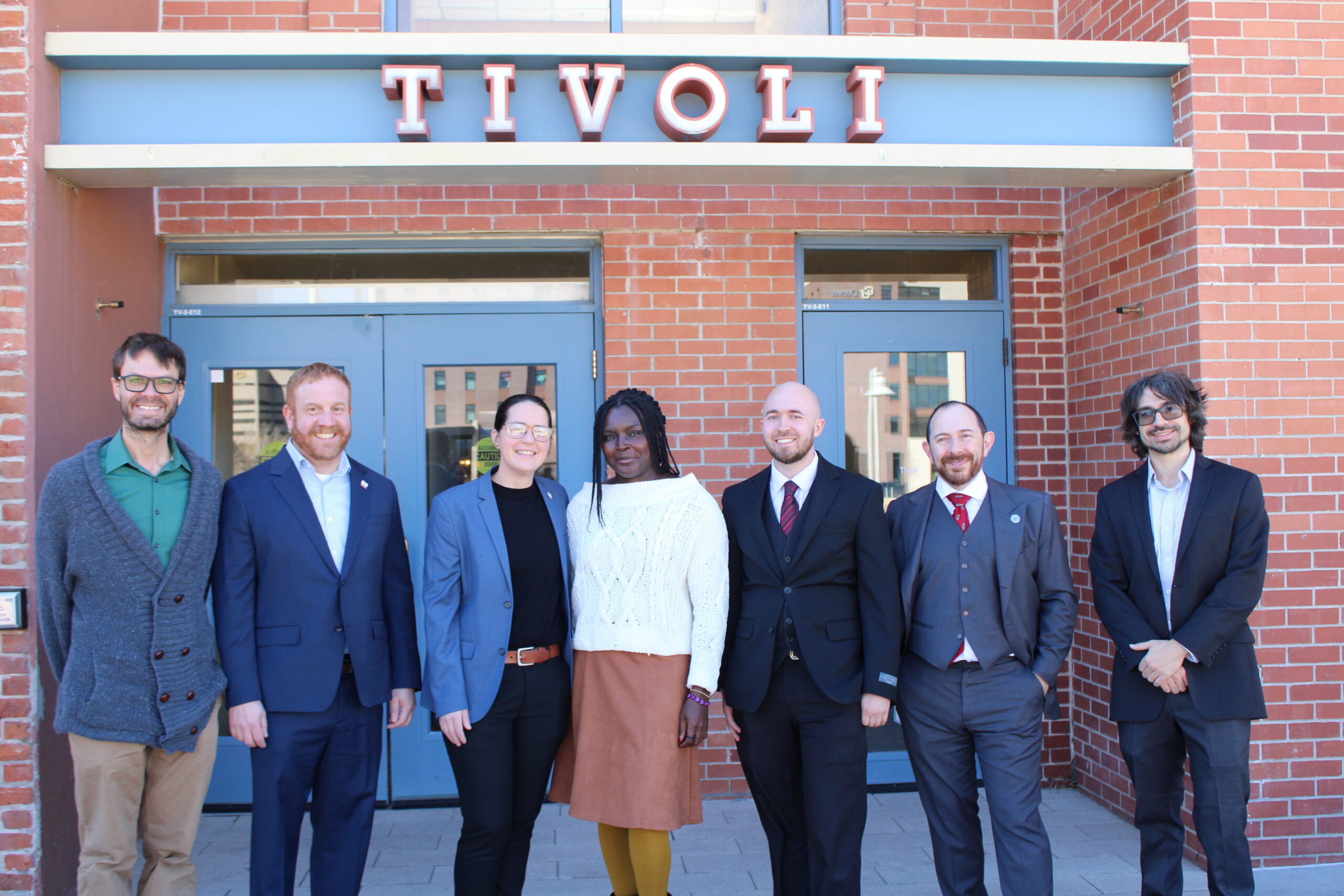 A group of seven professionally dressed people stand together outside in front of a brick building. They are positioned under a sign that reads 