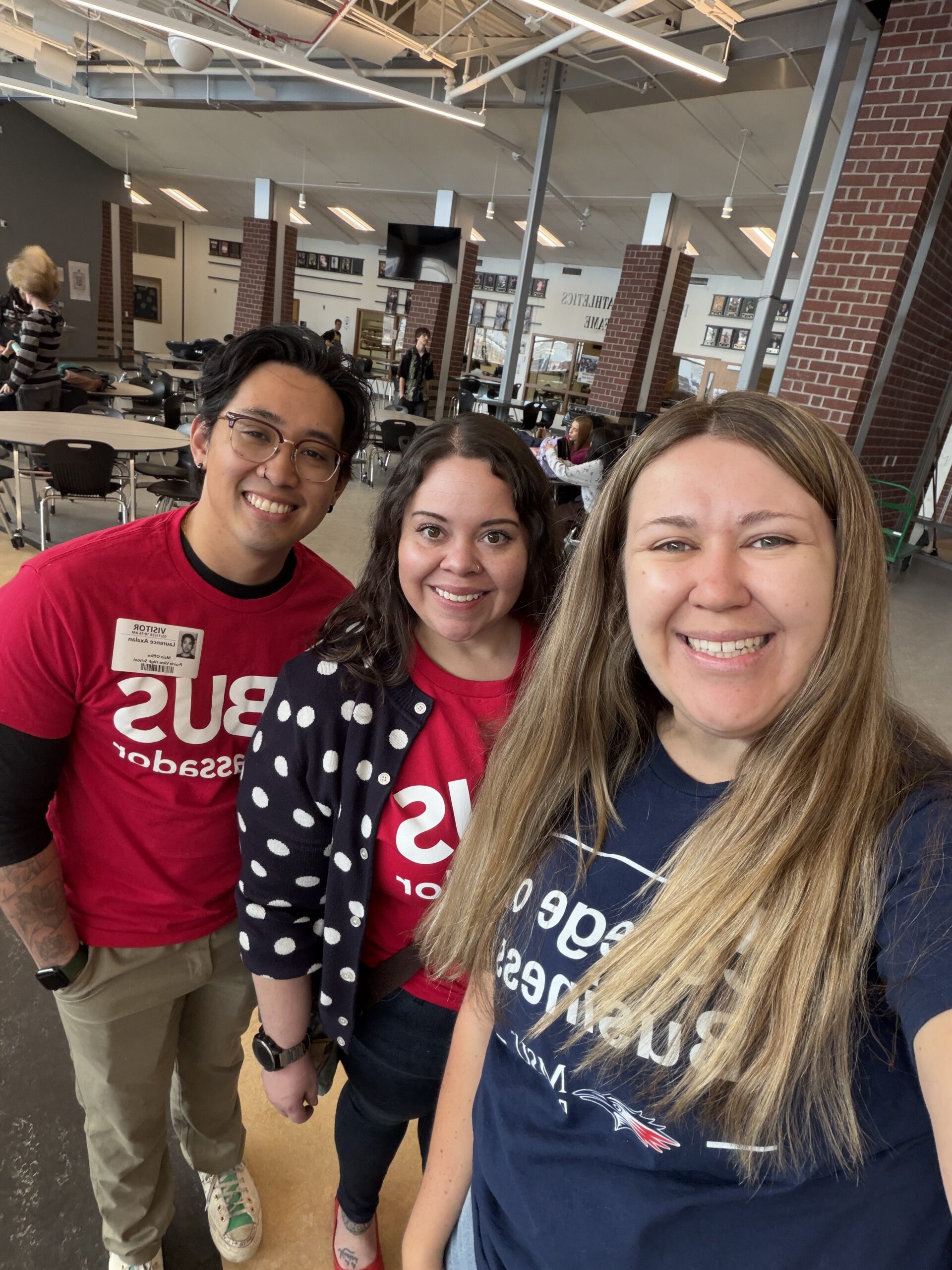 A high-angle selfie shows three people smiling in a large, open indoor space with high ceilings and brick pillars. The person on the left wears a red 
