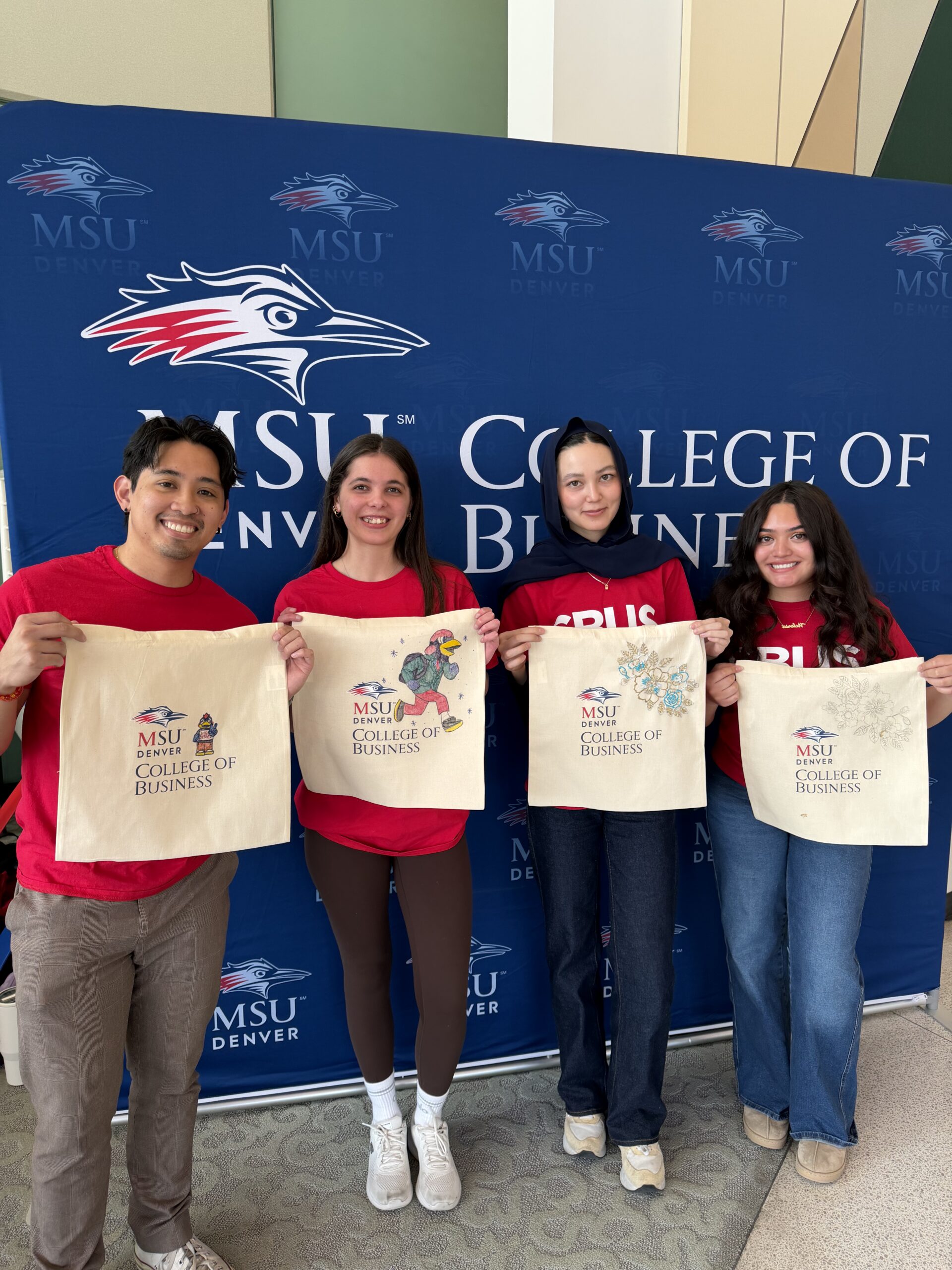 Four CBUS Ambassadors standing in front of the College of Business logo with red CBUS shirts holding their tote bags.
