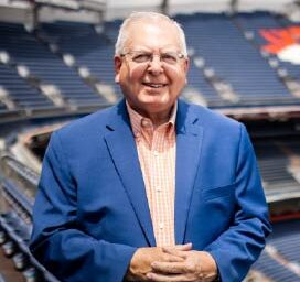 Man in a blue suit jacket and orange shirt stands with hands folded.