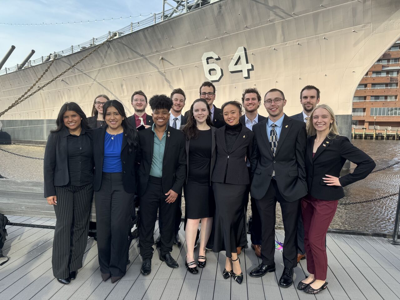 A group of twelve students in business casual and professional attire pose for a photo on a wooden pier. They are standing directly in front of the massive grey hull of a ship marked with the number 