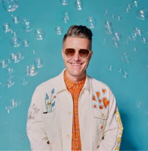 Man in a white jacket and sunglasses stands in front of a light blue background with bubbles