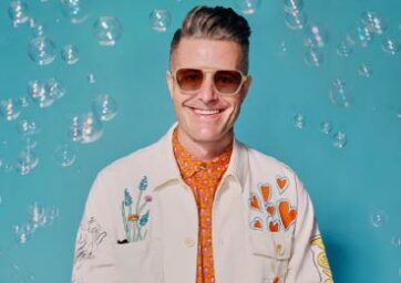 Man in a white jacket and sunglasses stands in front of a light blue background with bubbles