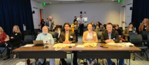 A panel of four judges: John Montaña, Luis Toscano, Chennelle Diong, and Aaron LaPedis—sit at a long wooden table during a business pitch competition at MSU Denver. They are smiling at the camera, with microphones and laptops positioned before them. The background shows a lively event space with attendees and organizers in a professional setting.