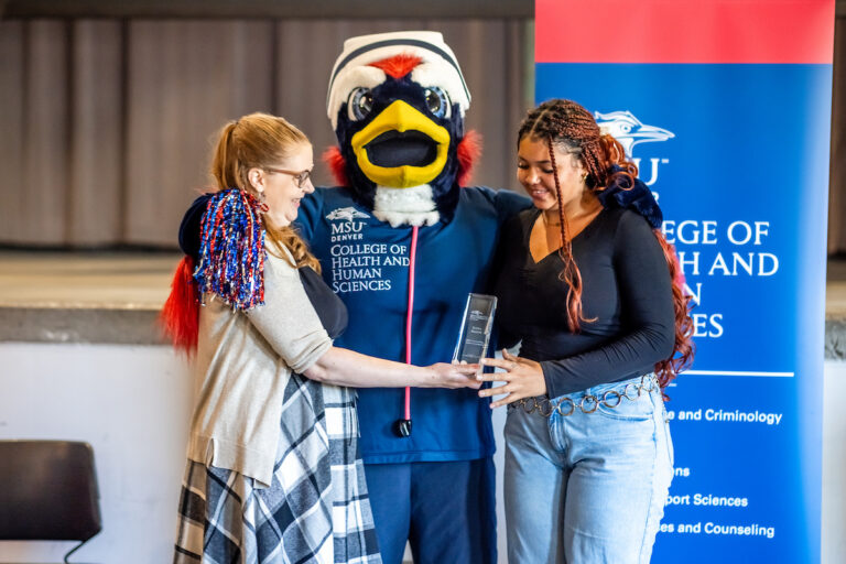 Caitlin Plamp (left) hands an Outstanding Student Employee Award to CHHS Peer Mentor Kayla Feldon (right) in the Tivoli Turnhalle on 9/24/25, while Nurse Rowdy (center) celebrates. Photo: Alyson McClaran