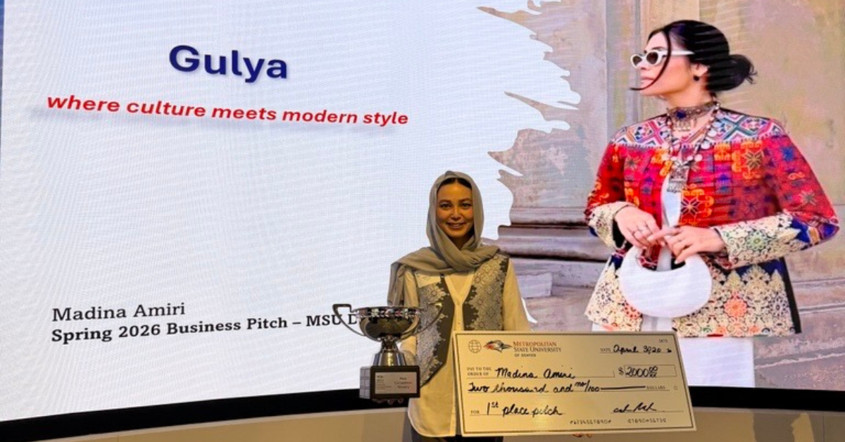 A woman named Madina Amiri stands proudly in a presentation hall, holding a trophy and a large ceremonial check after winning a business pitch competition. Behind her, a large digital screen displays her brand name, 