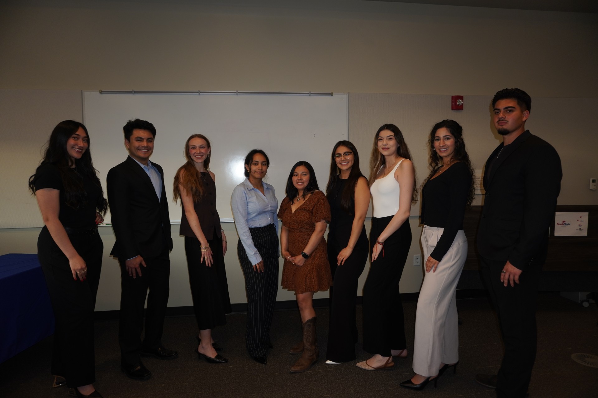 A group of nine young professionals from the Rowdy Minds Marketing Agency standing in a line and smiling in front of a white board. They are dressed in business casual attire, including suits, blouses, and slacks.