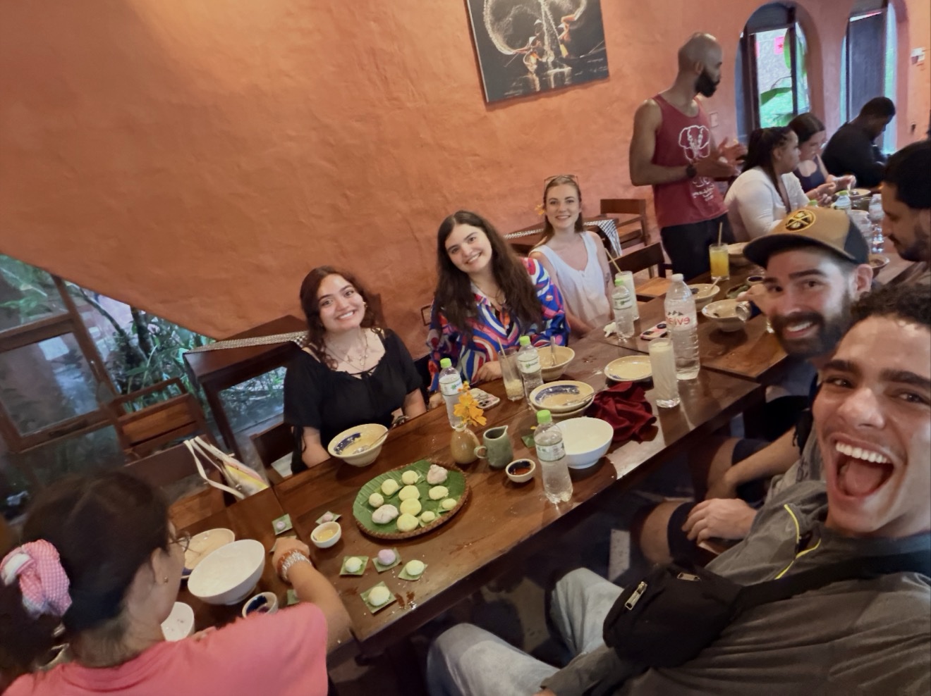 A high-angle group photo of students gathered around a long wooden dining table inside a restaurant with warm, textured orange walls. In the right foreground, a male student takes a selfie with an ecstatic, open-mouthed smile. Down the table, several other students smile towards the camera. The table is set with bowls, water bottles, and a central platter containing colorful, traditional round rice cakes or desserts served on a green leaf.