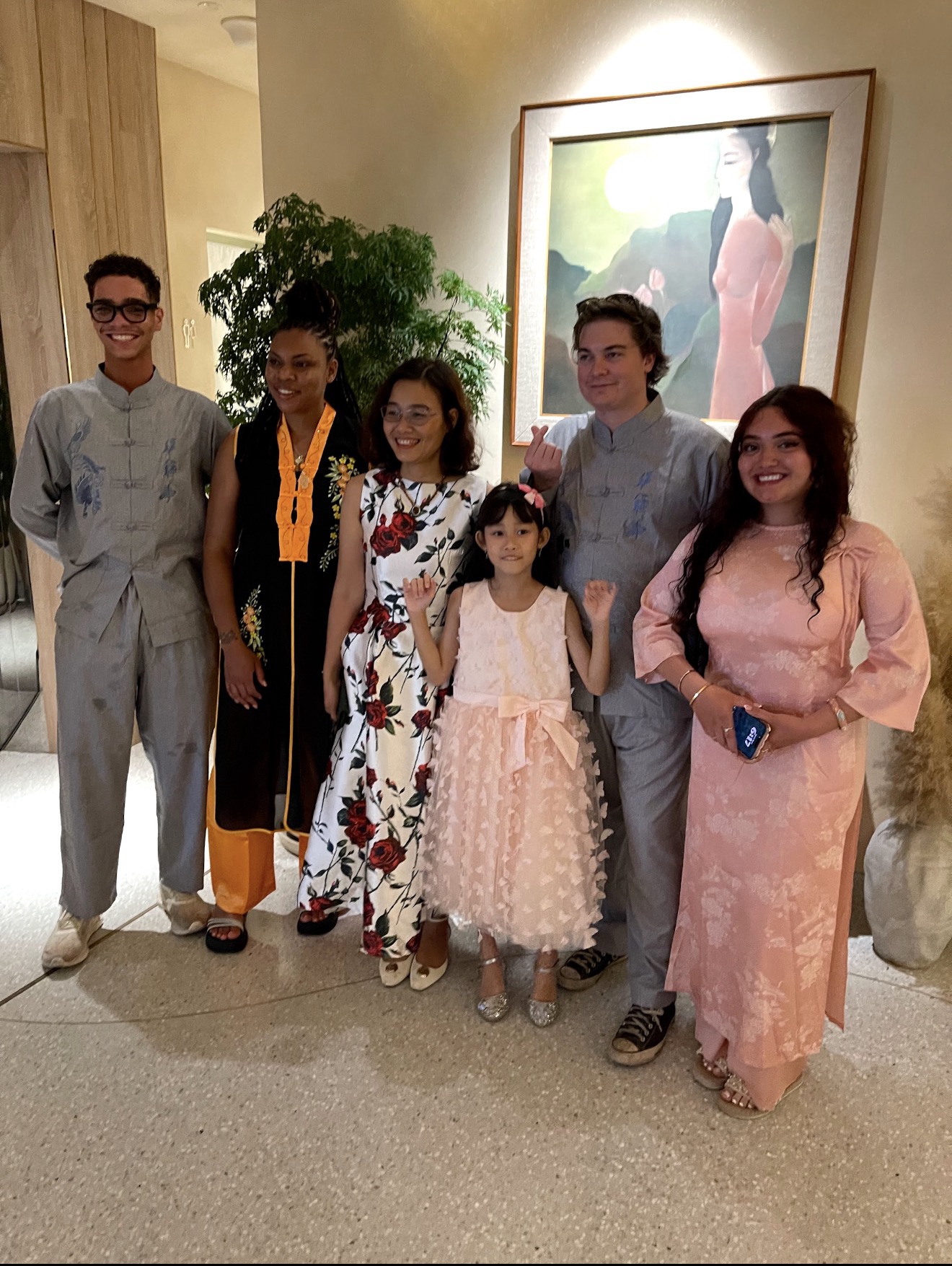 A group of six people posing for a photo indoors next to a painting of a woman in a landscape. Two of the male MSU Denver students wear matching grey, traditional-style frog-button shirts and trousers. Two women wear elegant, colorful traditional Vietnamese Ao Dai dresses—one in a white fabric with large red roses and the other in a soft pink patterned fabric. A young girl in a pink dress stands smiling in the center of the group.