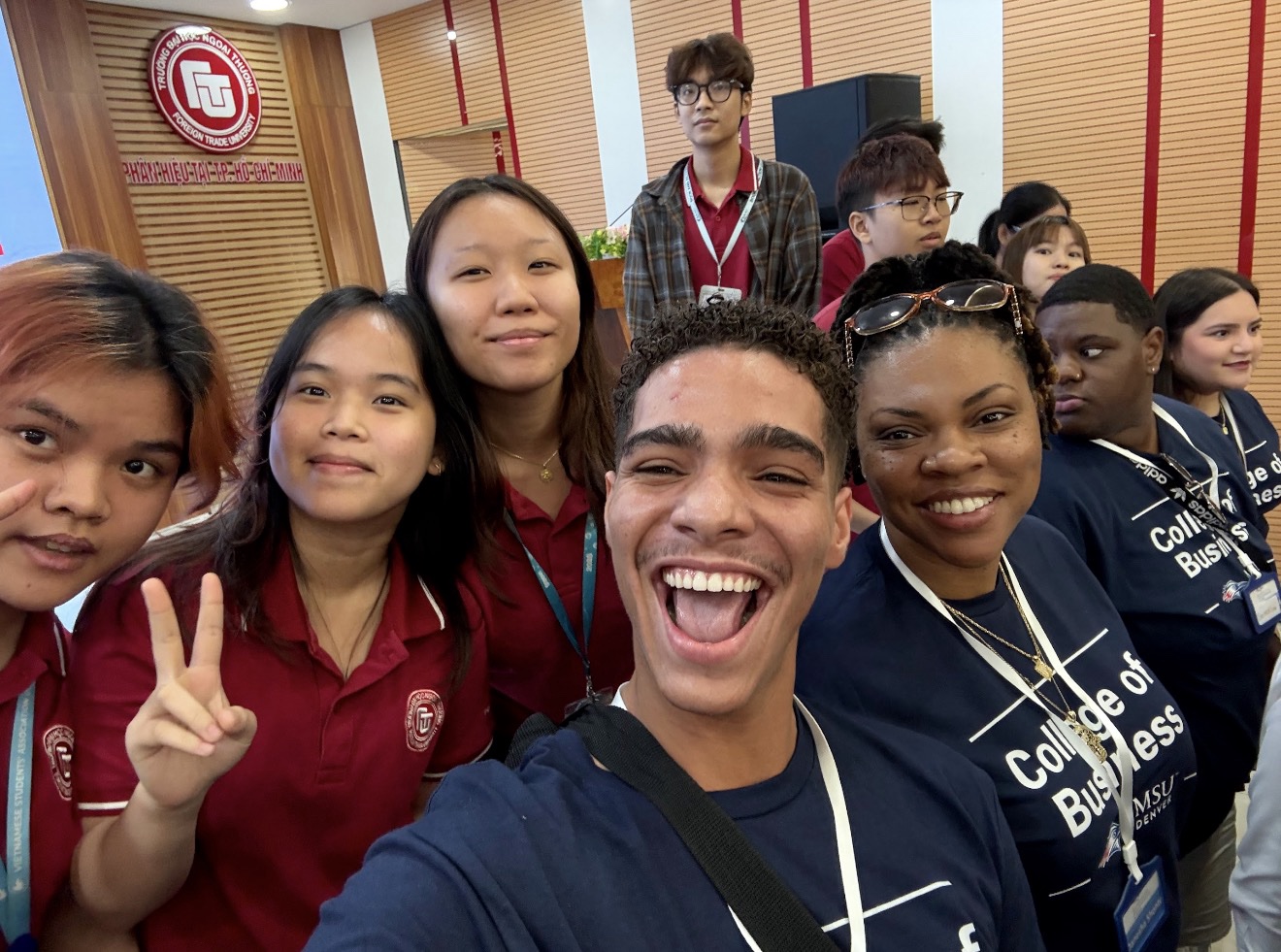A joyful group selfie featuring MSU Denver students and local Vietnamese students together at Foreign Trade University in Ho Chi Minh City. In the foreground, a male student smiles widely with an open-mouthed expression, while a woman beside him in a navy blue 