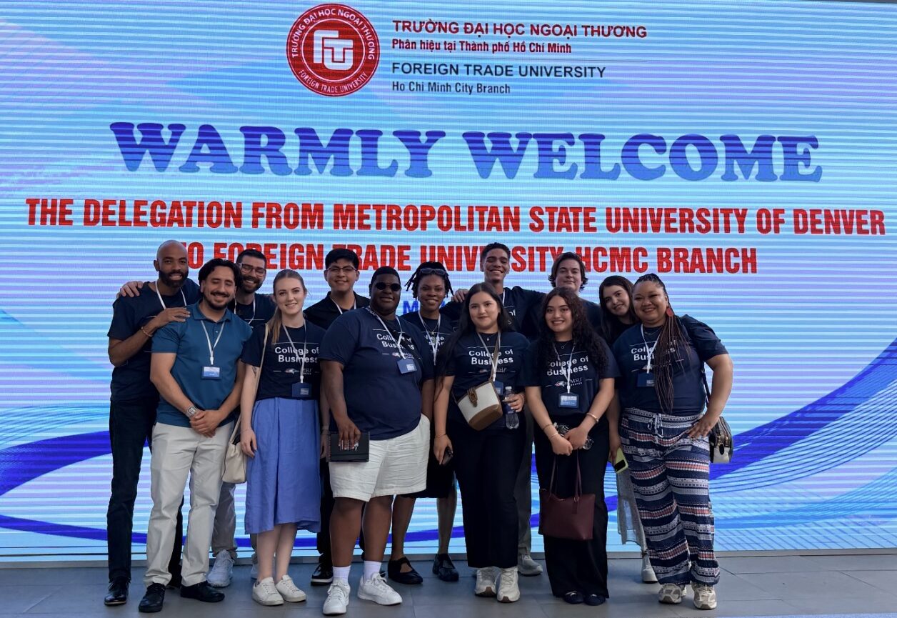 A group photo of the Metropolitan State University of Denver student delegation posing together on their study abroad trip. They are standing in front of a large digital screen that reads: 