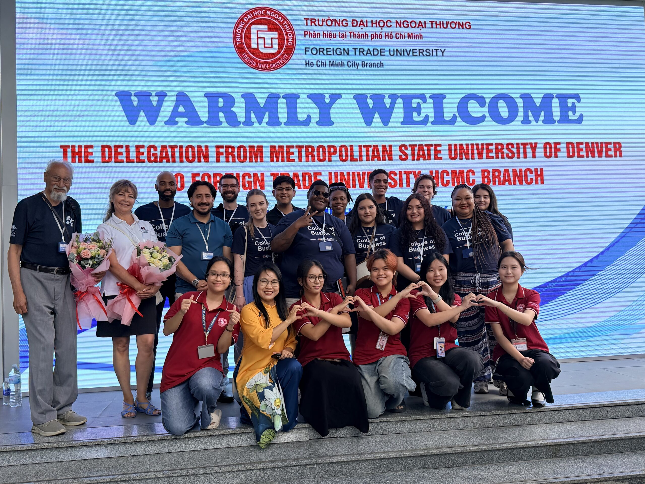 A large group photo of the MSU Denver delegation standing on a red carpet in front of a wide entryway arch. The arch is decorated with a blue graphic design and features the large Vietnamese word 