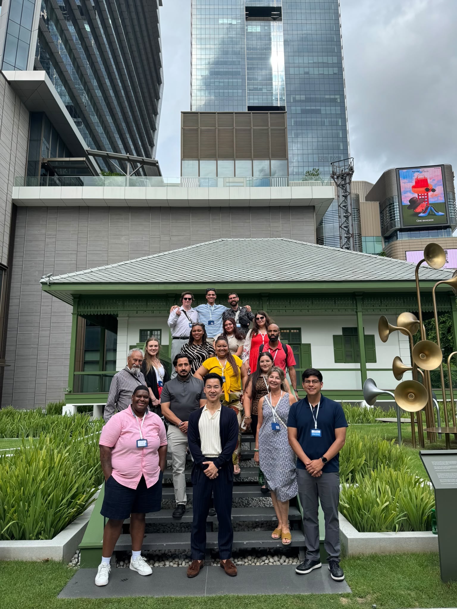 A group photo of the MSU Denver delegation arranged on a set of outdoor steps in front of a traditional, white-and-green single-story house. Strikingly contrasted directly behind this traditional building is a massive, modern glass skyscraper rising high into the sky. Lush green grass and modern horn-shaped sculptures flank the steps.