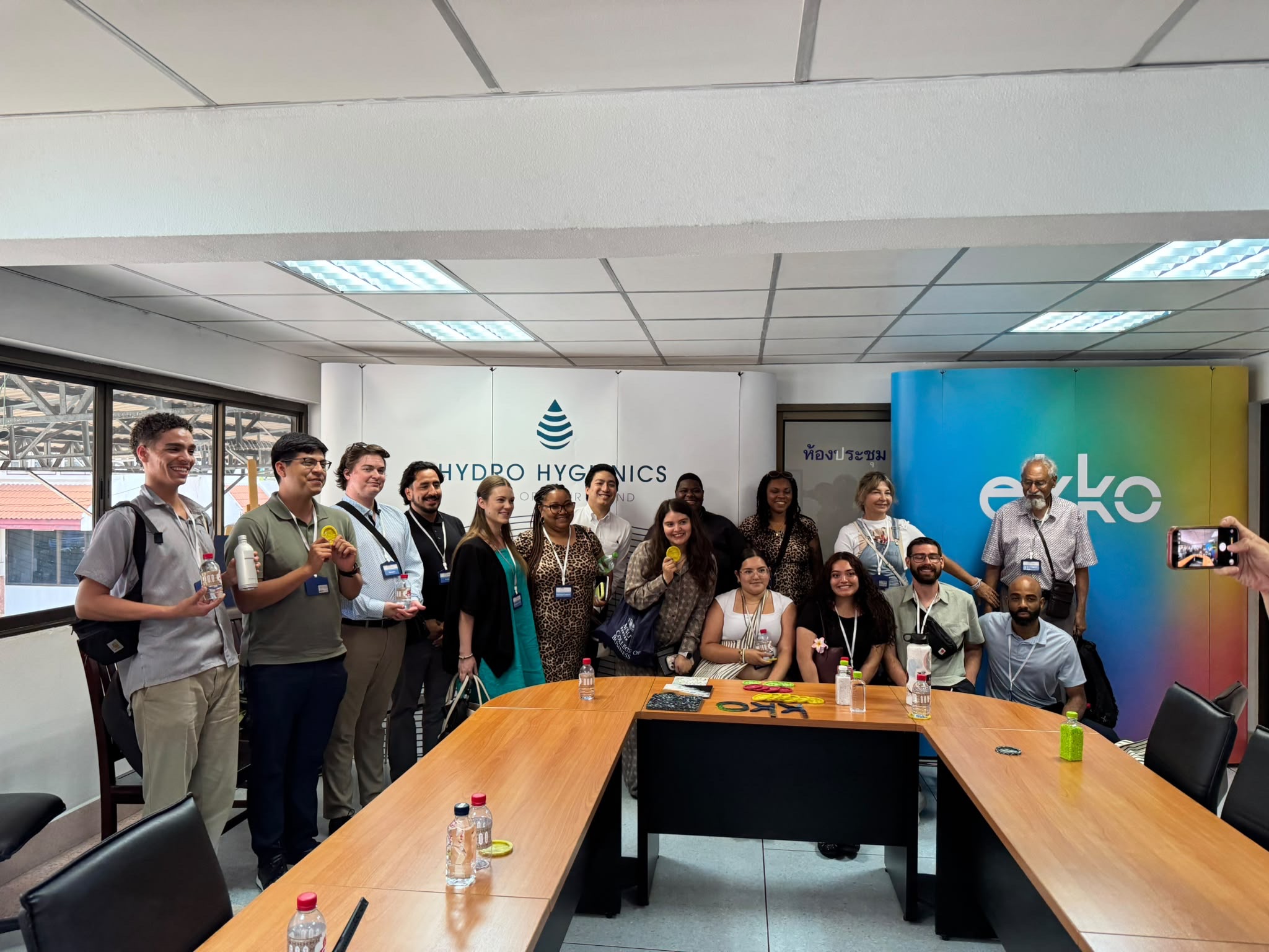 The student delegation posing around a large wooden, U-shaped conference table inside a corporate meeting room. Behind the group is a white backdrop that reads 