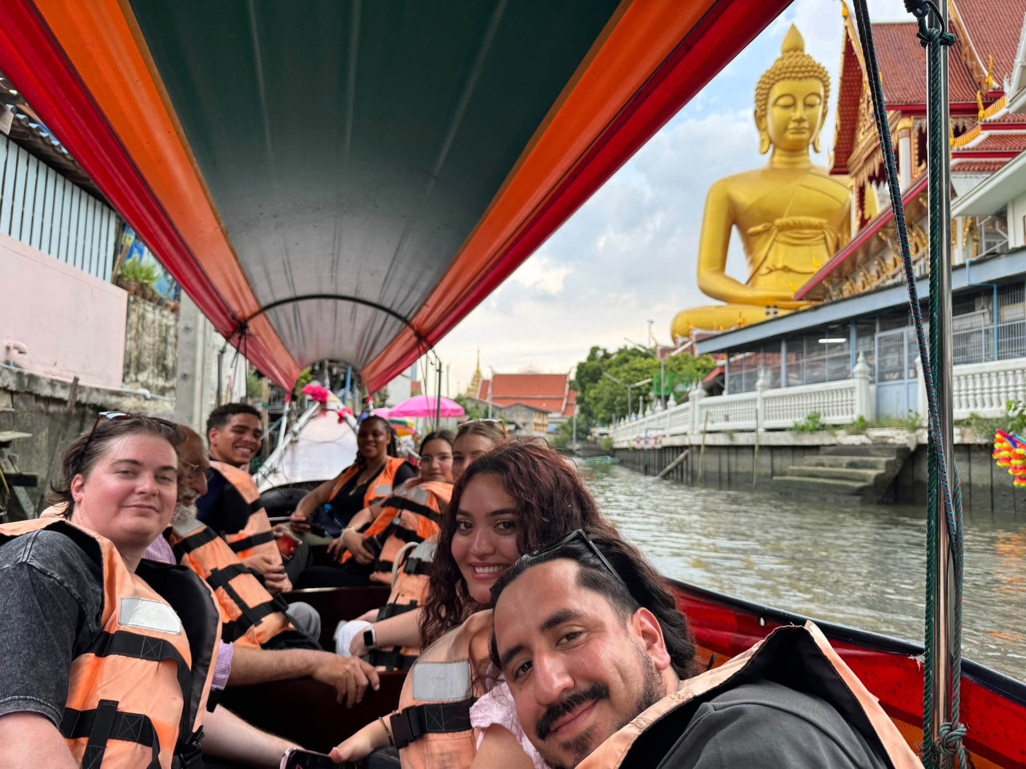 A group selfie taken from the front of a long, narrow canopy boat traveling down a canal in Thailand. In the immediate foreground, a male student and a female student smile at the camera. In the background on the right bank, a massive, towering golden statue of a seated Buddha rises above the nearby buildings under an overcast sky.