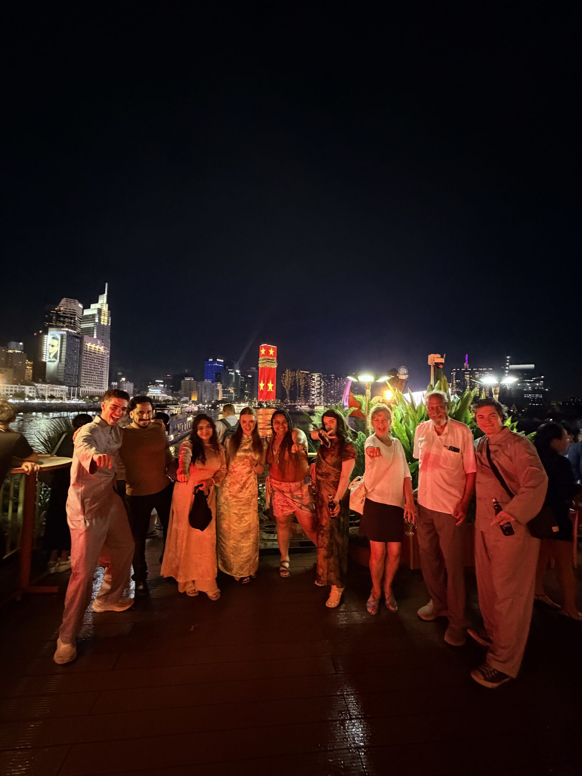 A vibrant nighttime group photo of the students posing together on a beautifully lit outdoor terrace or rooftop. The area is decorated with lush hanging plants, warm string lights, and glowing spherical lanterns, with city buildings visible in the background under the dark sky.