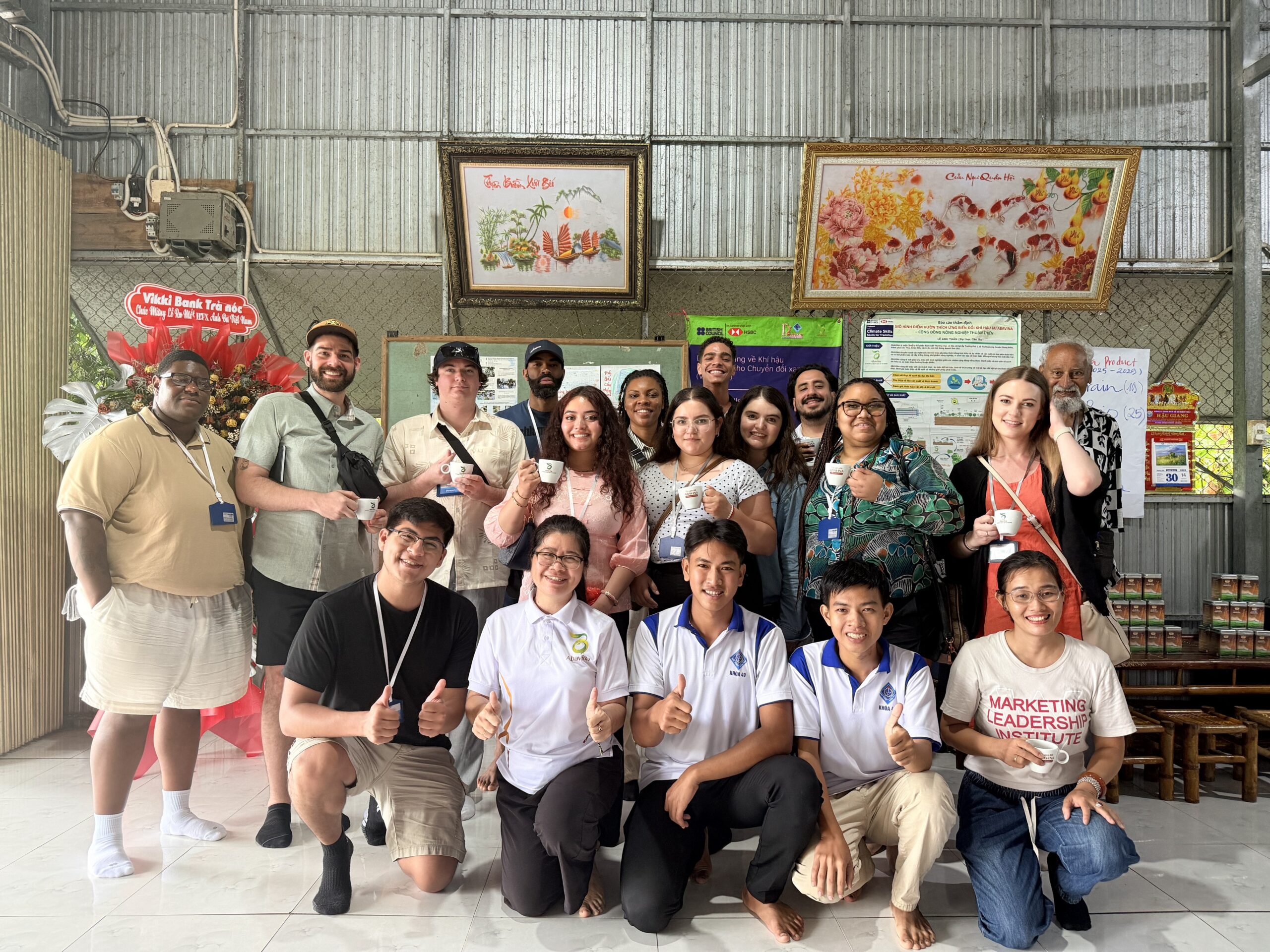 A group photo of MSU Denver students posing indoors alongside local Vietnamese hosts or students. They are gathered in a room with corrugated metal walls, and many individuals are holding up small white espresso or tea cups, smiling toward the camera. Two framed pieces of art hang on the wall behind them.