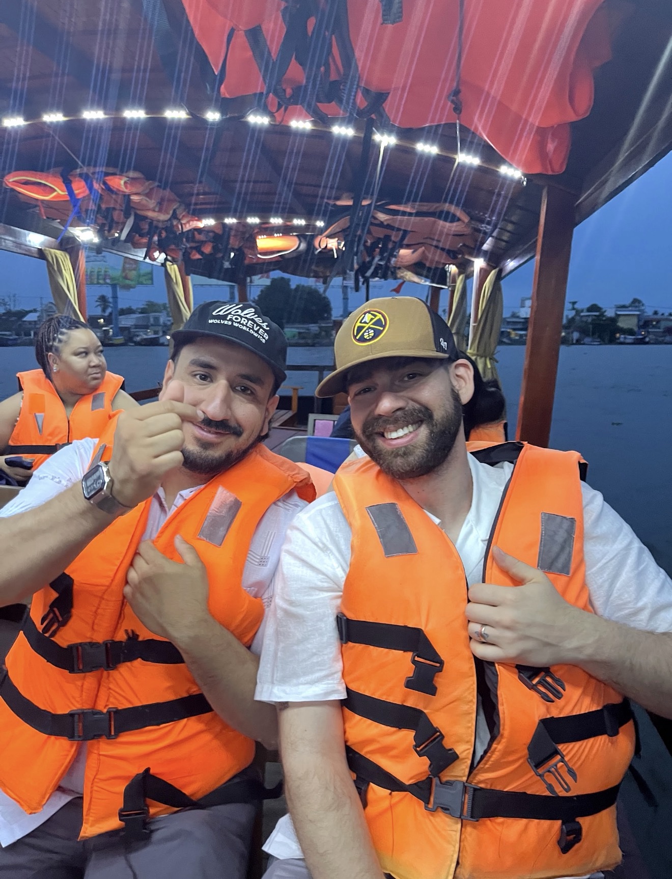 Two male MSU Denver students sitting closely together on a boat cruise at dusk, both wearing bright orange life jackets. The student on the left wears a black baseball cap and makes a small finger-heart gesture with his hand, while the student on the right wears a brown baseball cap and smiles broadly at the camera. Other passengers and life jackets are visible under the boat's canopy behind them.