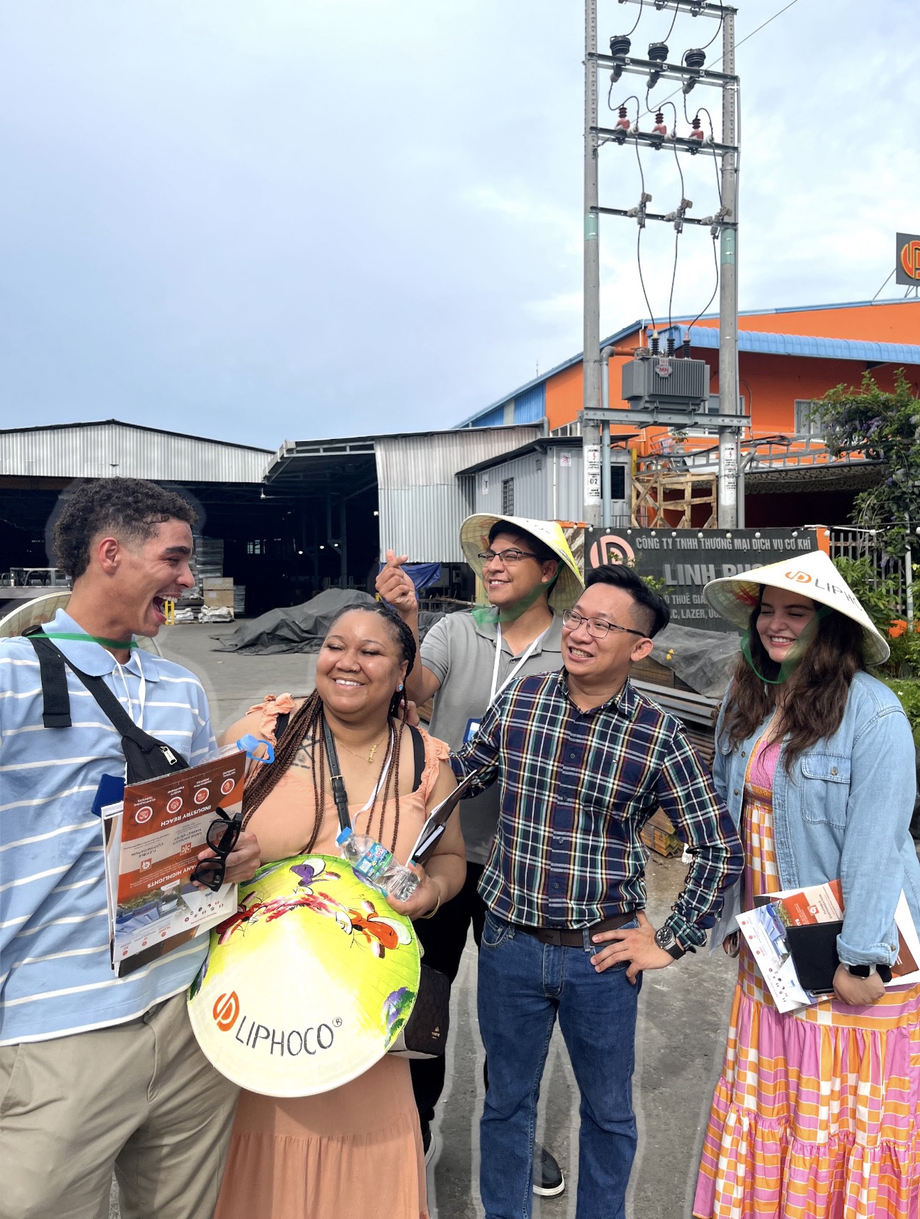 Five people standing outside an industrial or commercial building, laughing and smiling together. Three individuals are wearing traditional conical hats, and one male student on the left is holding a brochure while laughing with an open mouth. A local representative in a plaid shirt stands in the center with his hand on his hip, smiling. An electrical pole with transformers stands behind them against a cloudy sky.
