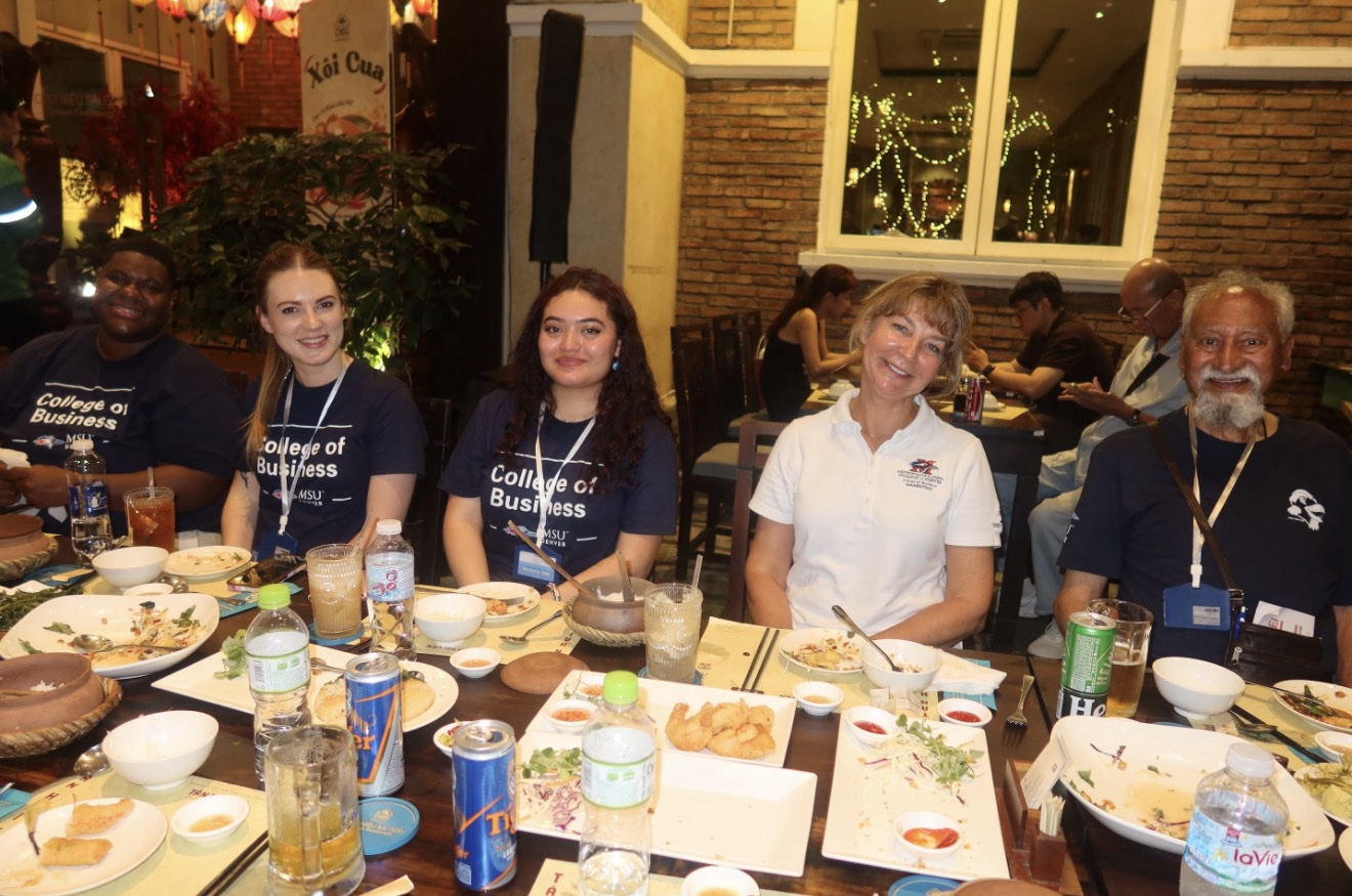 A group of MSU Denver students and faculty sitting along one side of a long wooden dining table during a group meal at a restaurant in Vietnam. Some students are wearing matching navy blue 