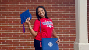 Stacy Berry standing in front of a brick wall holding her diploma and graduation cap
