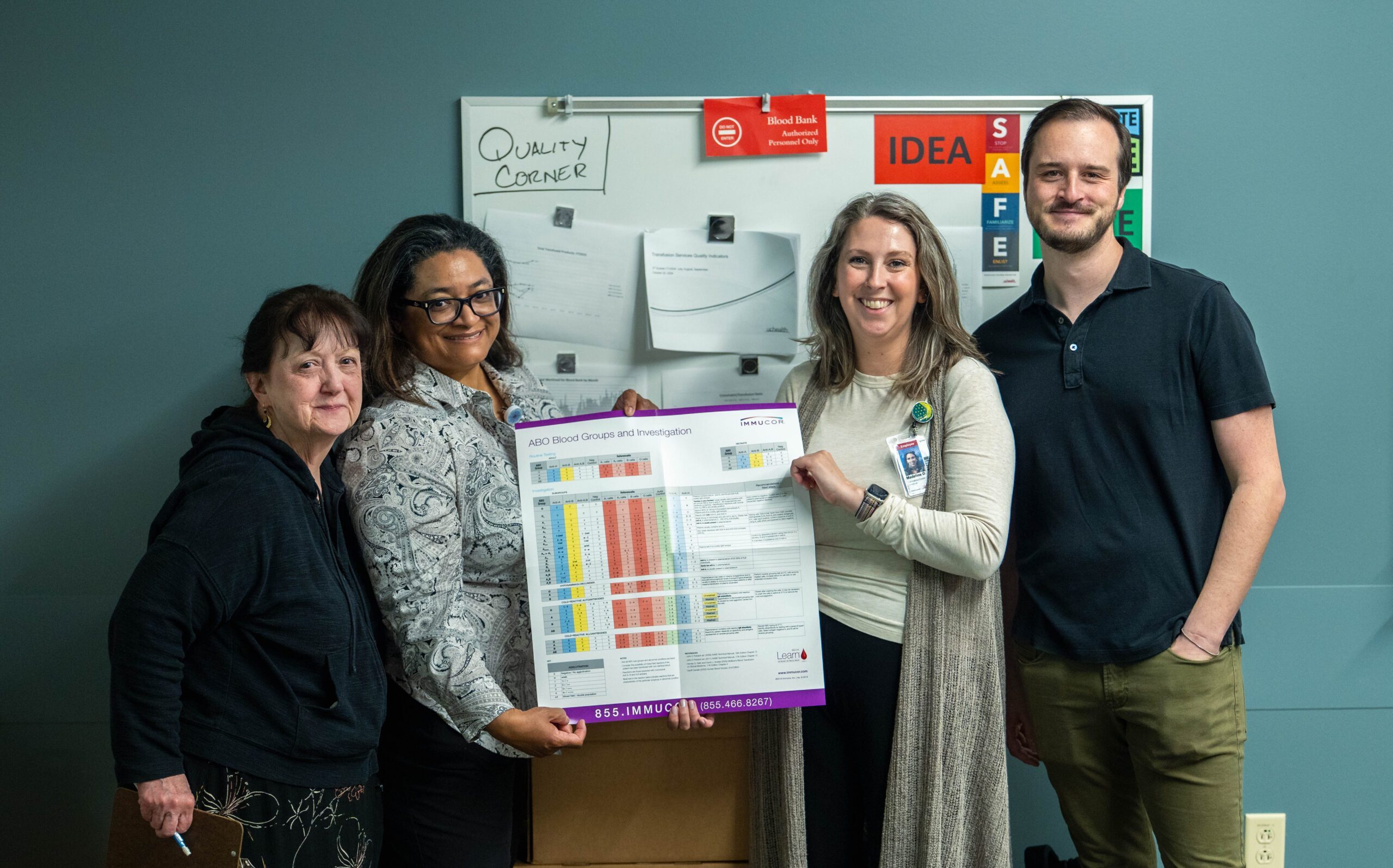 A group of four holding up a poster in front of a whiteboard and colored wall.