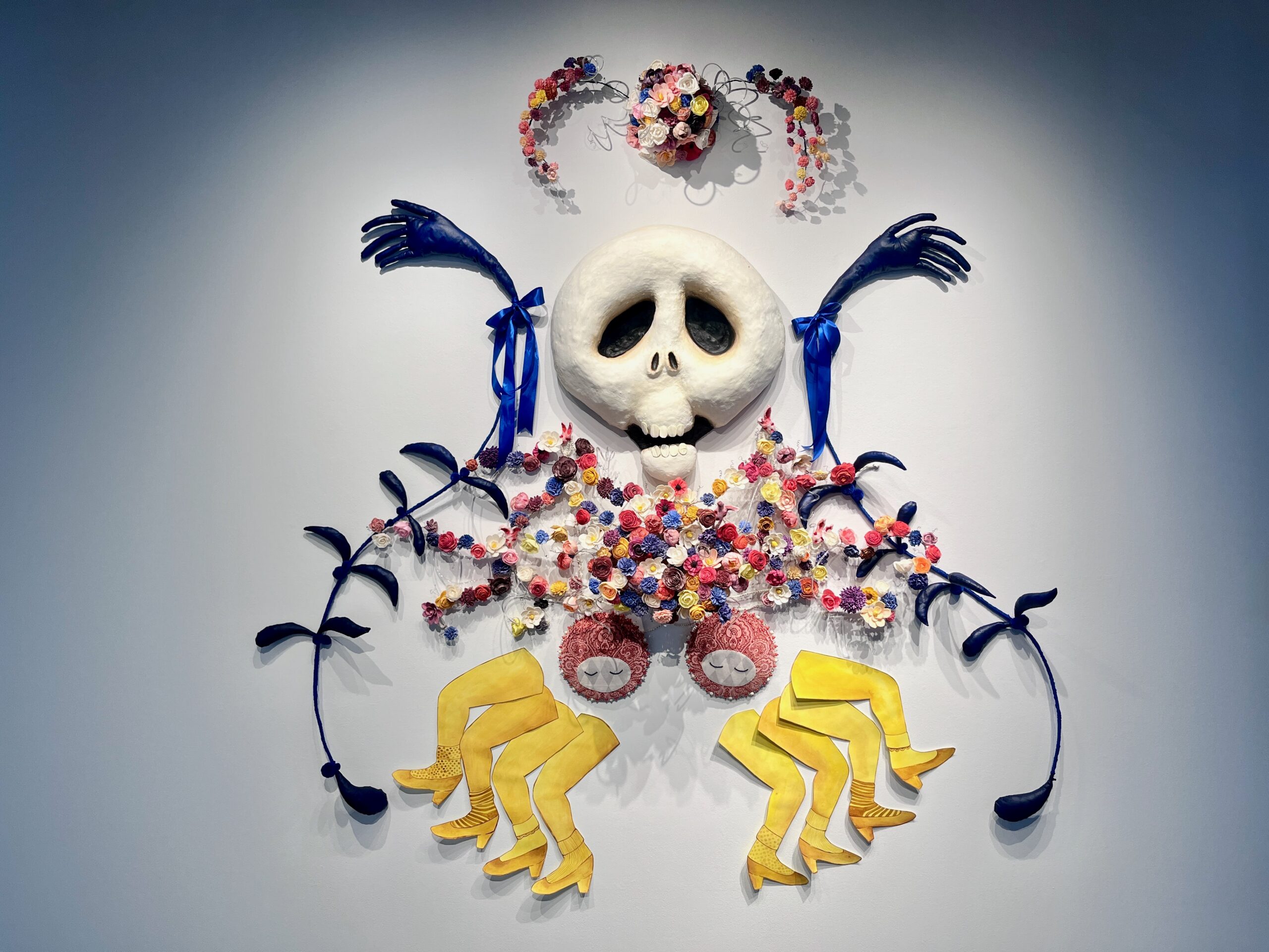 A wall installation featuring a large white skull surrounded by colorful artificial flowers, blue hands with ribbons, and whimsical yellow legs.