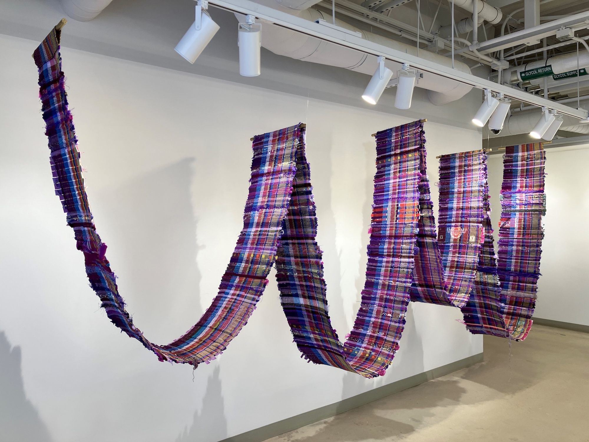 A large textile installation featuring colorful, wave-like strips of fabric in shades of purple, pink, and blue, suspended in an art gallery.