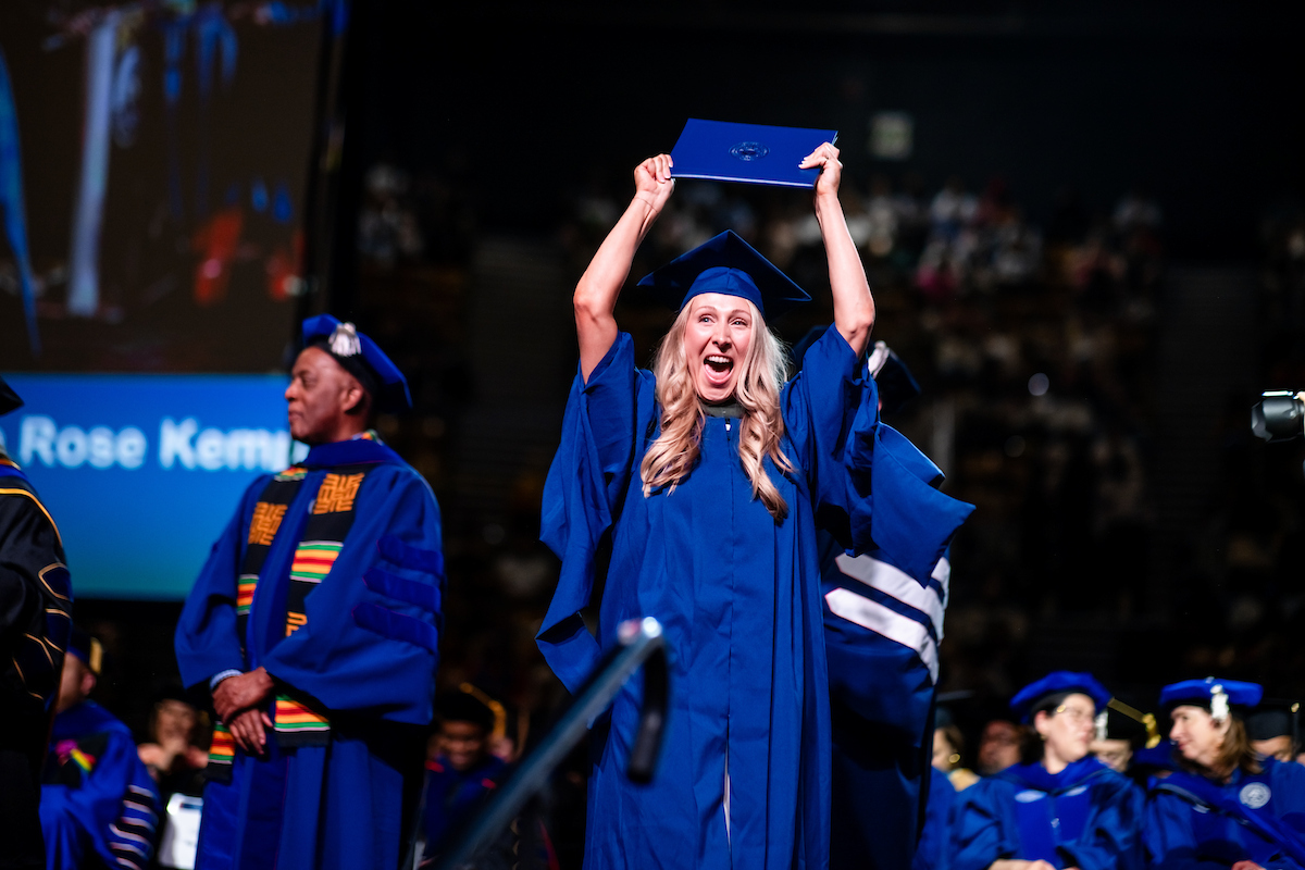 MS SLP graduate in a blue cap and gown stands on a commencement stage holding a diploma cover overhead while receiving recognition. Faculty members in academic regalia are seated and standing nearby, with a large screen and audience visible in the background.