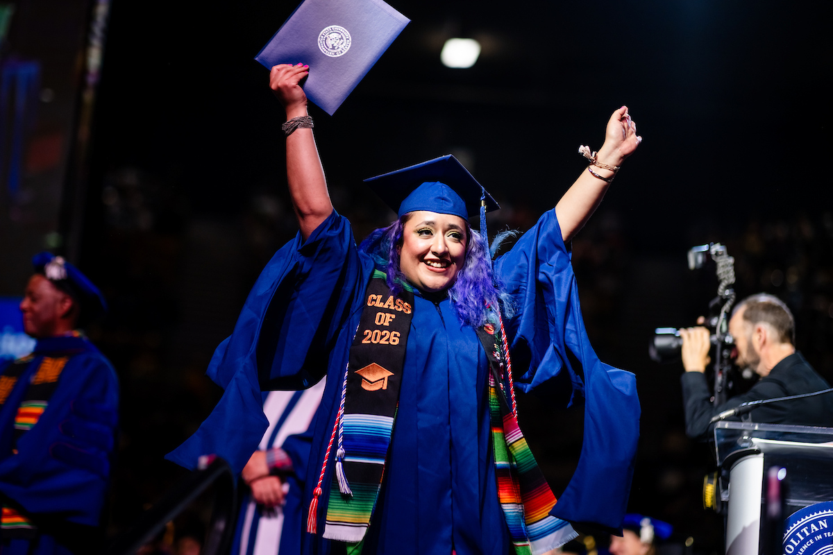 MS SLP graduate in a blue cap and gown raises a diploma cover overhead while walking across the commencement stage. The MS SLP graduate wears a black stole with the visible text “Class of 2026,” a multicolored stole, and honor cords. Faculty members in academic regalia, a photographer, and a podium are visible in the background.