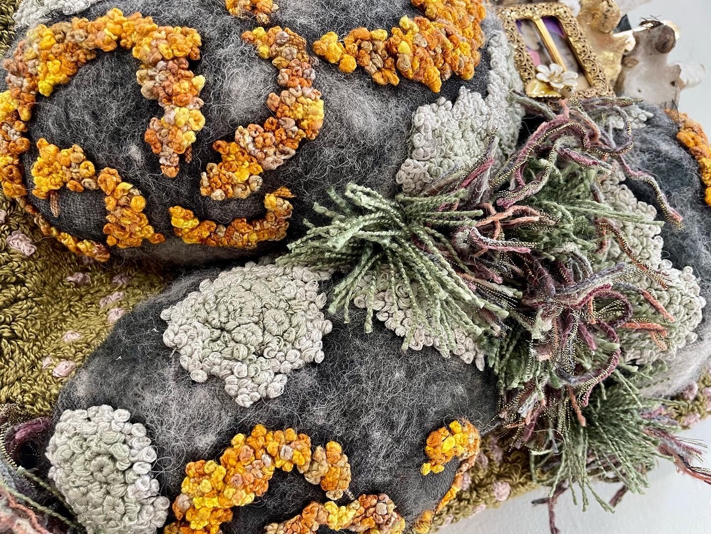 A close-up of an artistic textile piece showcasing dark, textured fabric with vibrant orange and yellow embroidery, resembling a lichen covered rock, complemented by green and grey stitches.