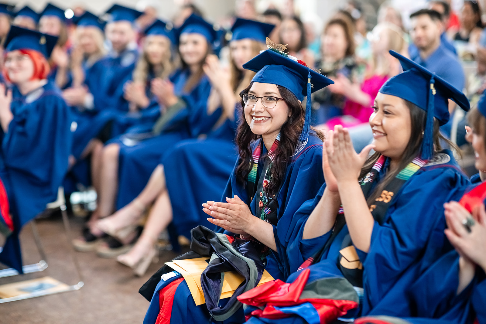 MS SLP graduates in blue caps and gowns sit together during a hooding ceremony and applaud while facing toward the stage. Two MS SLP graduates in the foreground wear decorative honor stoles and academic regalia, with folders and ceremonial hoods resting on their laps. Rows of MS SLP graduates and guests are visible in the background of the indoor venue.
