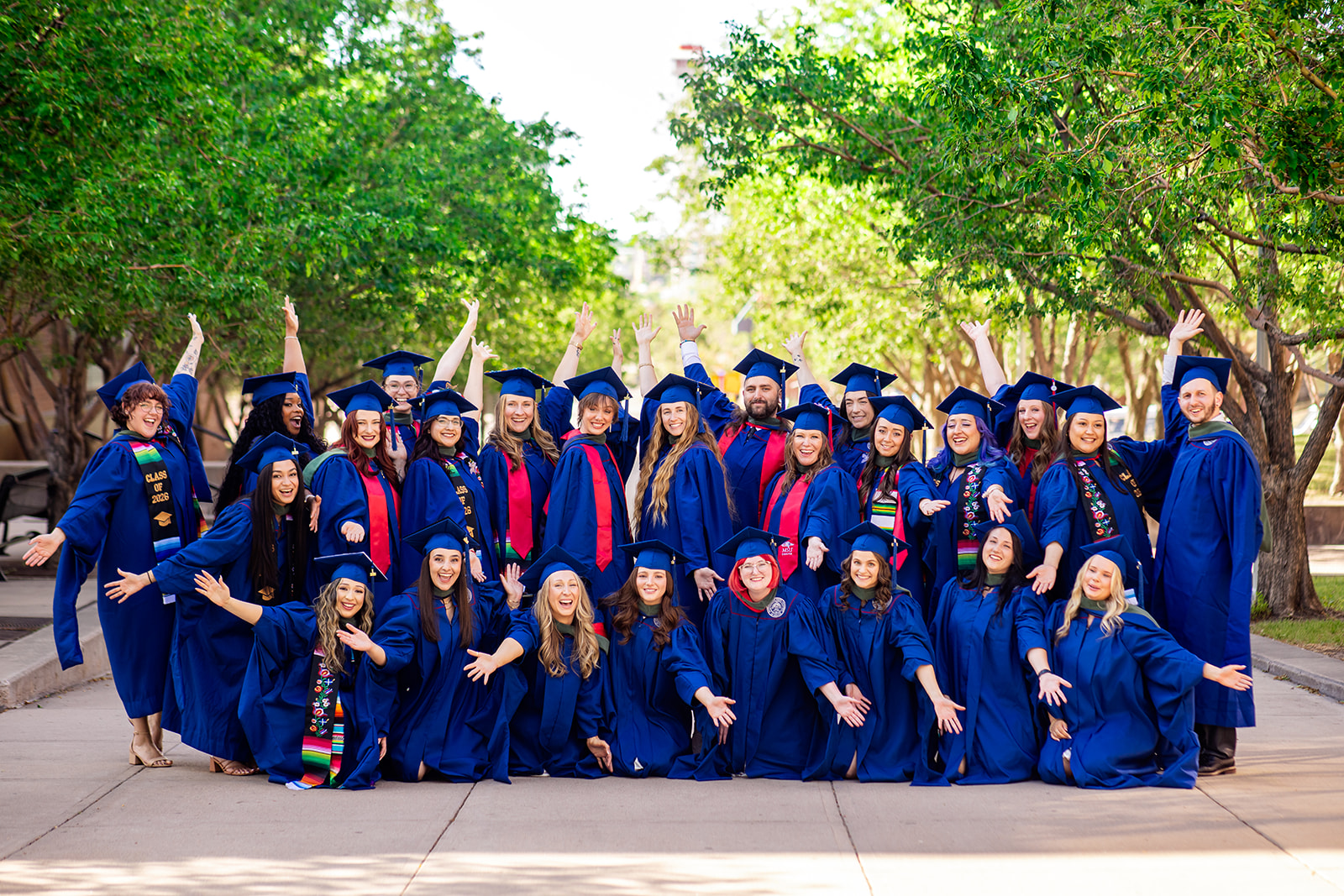 A large group of MS SLP graduates in blue caps and gowns pose together on a tree-lined campus walkway during a graduation celebration. Several MS SLP graduates wear red academic stoles, colorful honor stoles, and ceremonial hoods. The group is arranged in standing and kneeling rows with arms raised and extended toward the camera, while green trees and campus buildings frame the outdoor setting.