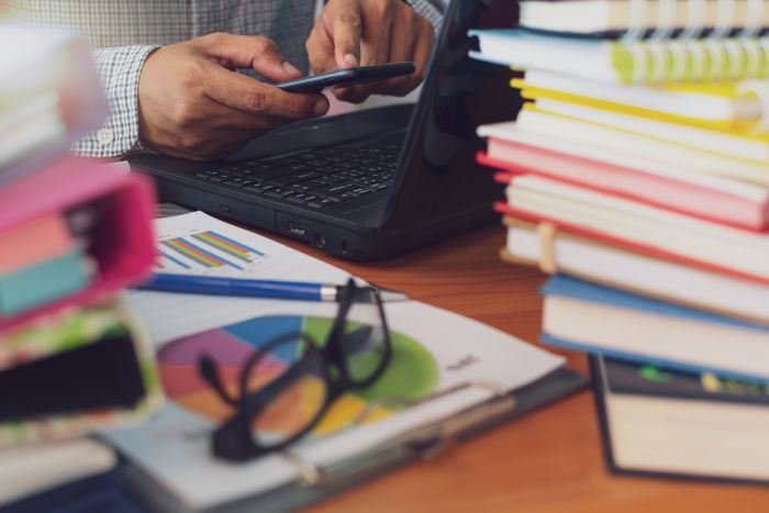 An image of a desk covered in books and papers with someone on their cell phone in front of a laptop