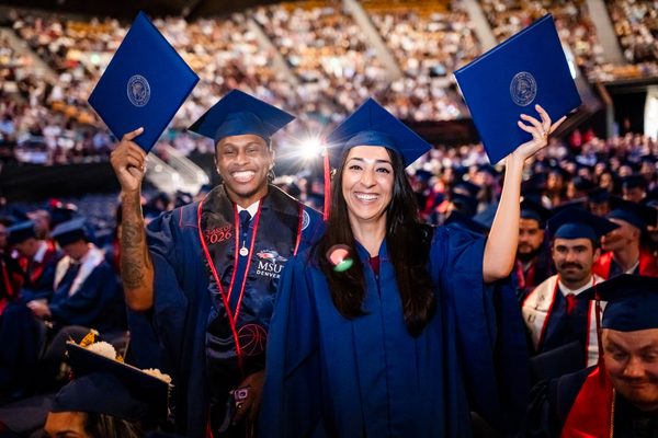 Two grads smile and pose for the camera, shoeing off their new diploma covers