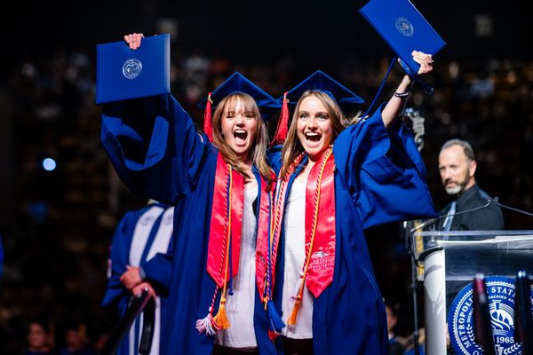 Two grads pose with their diploma covers victoriously and smile for the camera