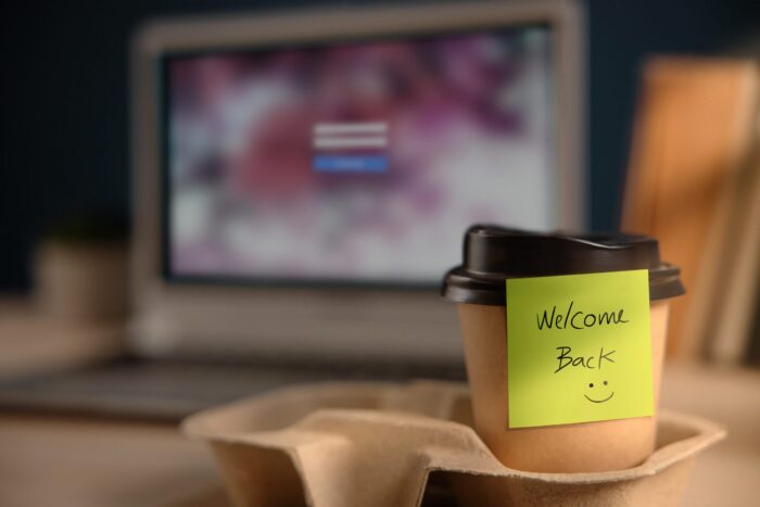 A takeaway cup of coffee with a Welcome Back note on. There is a blurred desk and computer in the background.