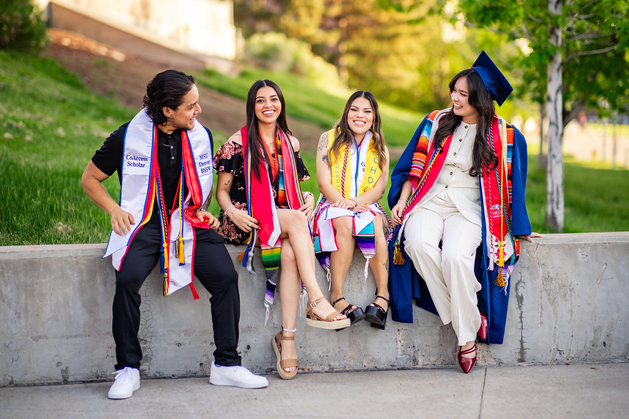 Four smiling BSSW MSU Denver graduates in colorful stoles and caps sit together on a campus wall, chatting and laughing in a sunny, green setting after graduation.