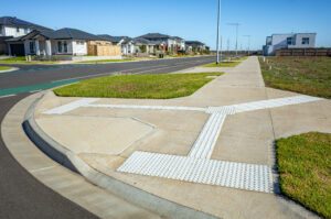 A newly constructed suburban sidewalk featuring tactile ground surface indicators (TGSIs) for visually impaired pedestrians at a curb ramp.The footpath runs alongside a residential street in Australia 