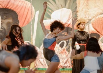 Curly-haired dancer in blue-red leotard raises arms center stage, other dancers and fabric mural backdrop behind.