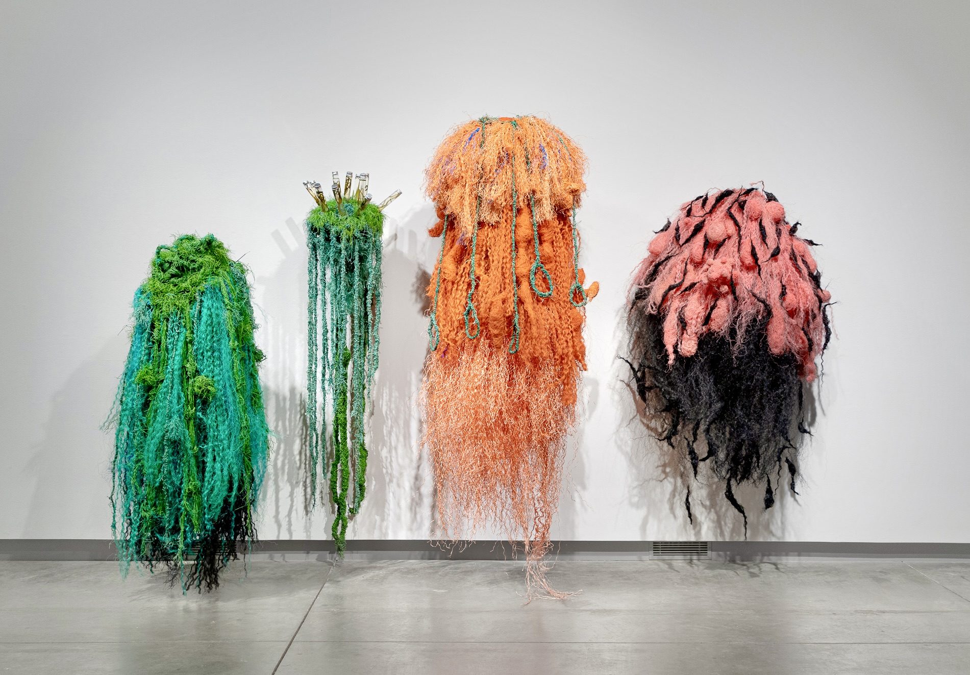 Four colorful, shaggy textile sculptures hang against a white gallery wall, in turquoise and green, green, orange, and pink and black.