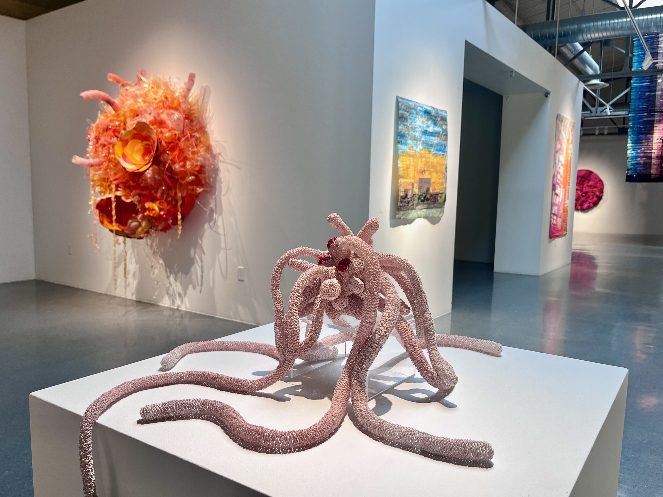 Contemporary gallery with a pale pink tentacle-like sculpture on a white pedestal and a glowing orange circular sculpture on the wall behind it.