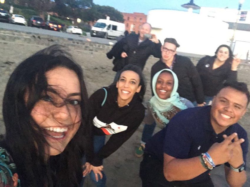 Six Model UN students in 2019, smiling in a group selfie on a beach at dusk while Richard Moeller takes a photo behind them.