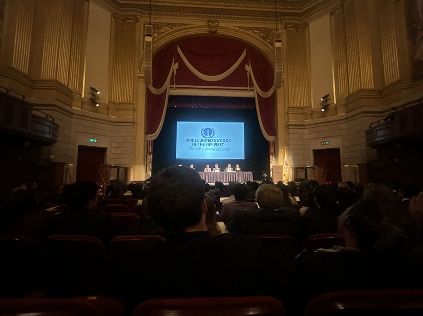 Model UN delegates in 2026, seated in an ornate auditorium facing a panel on stage beneath a screen reading 