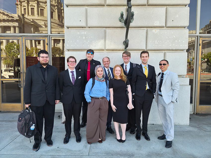 Richard Moeller and nine Model UN students in 2026, dressed in business attire and posing on a sidewalk in front of a stone building.