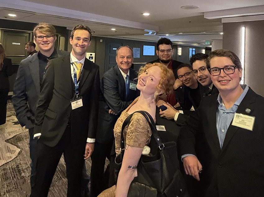 Richard Moeller and seven Model UN students in 2026, smiling together in a hotel hallway and wearing conference name badges.
