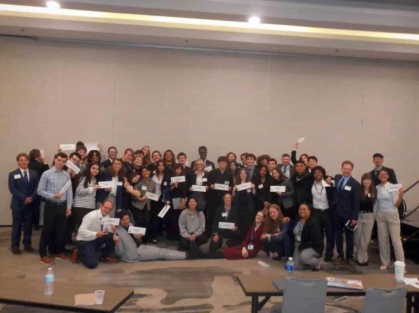 A large group of Model UN delegates in 2026, posing together in a conference room and holding up placards.