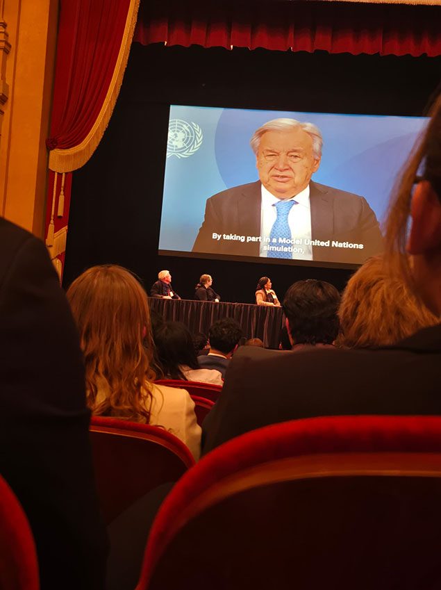 Model UN delegates in 2026, seated in an auditorium watching a video address on a screen showing the United Nations emblem, captioned 